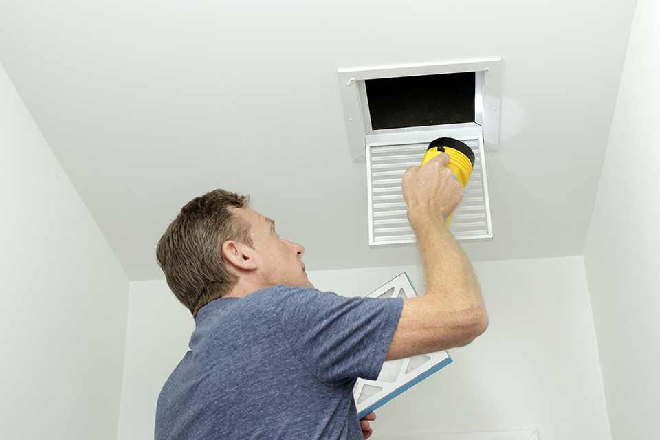 Man inspecting air ducts shining a flashlight through a small square ceiling vent into ducting pipes. Mature male examining the condition of air ducts at home.